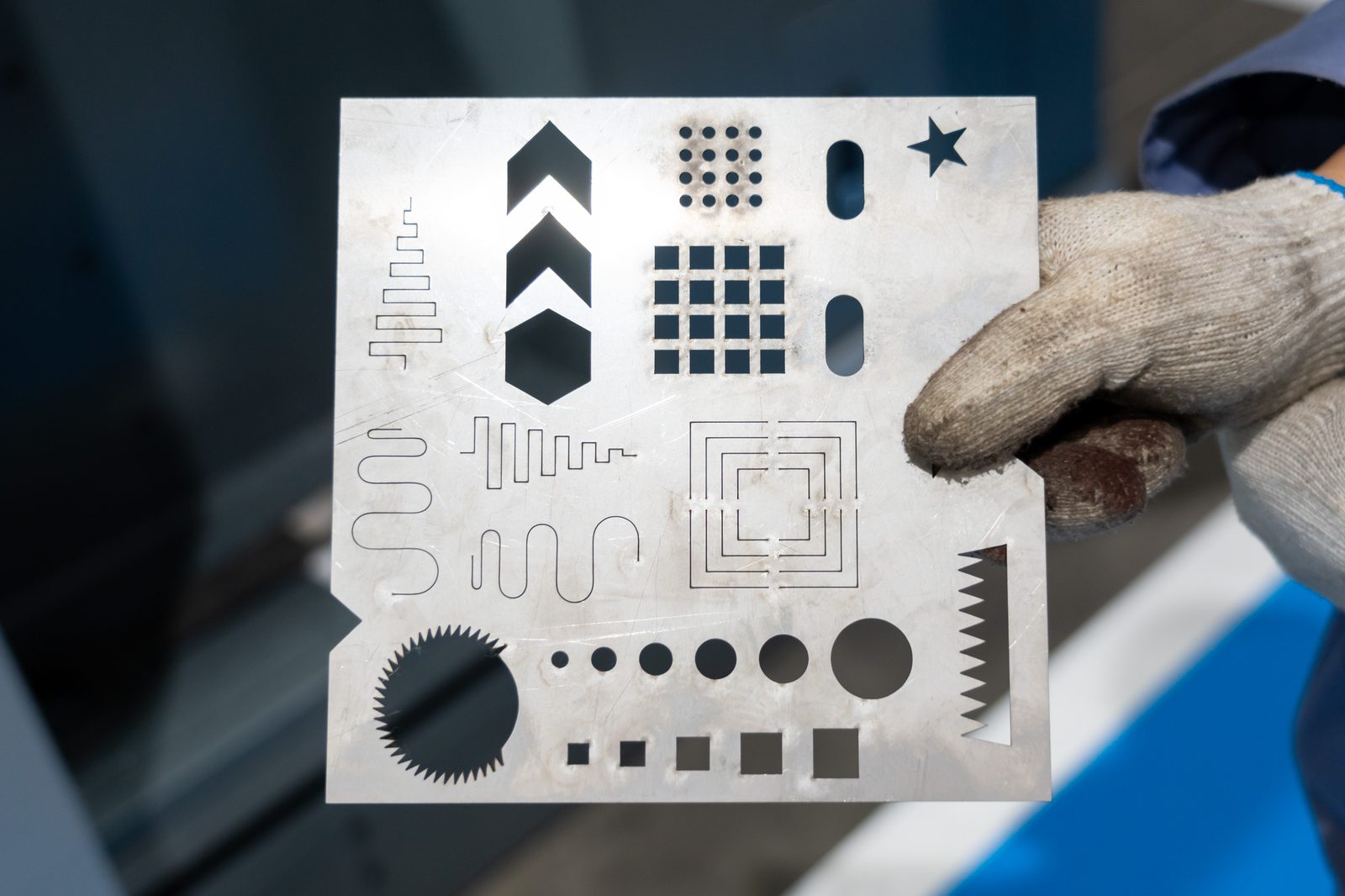 A gloved hand holding a laser-cut sample plate carrying gears, chevrons, slots and a graduated hole pattern